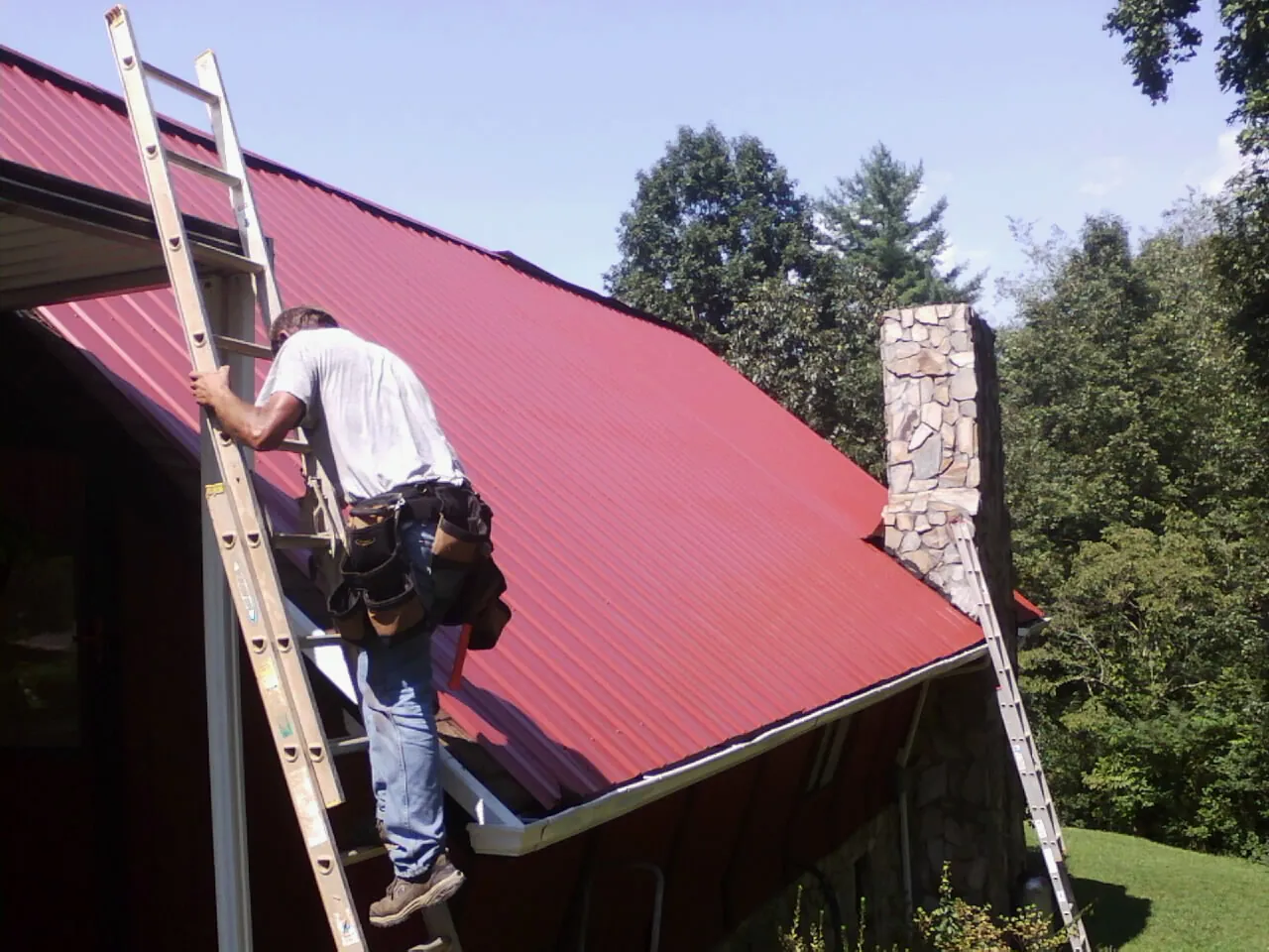 Roofer on a ladder beside a red metal roof and stone chimney — Soffit & Fascia Repair by Roofing Brothers in Humboldt